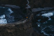 Сверху сзади вид человека с вытянутыми руками, наслаждающегося свободой, стоящего на старом каменном мосту против беспокойной воды с пеной, омывающей скалистые берега Испании — стоковое фото