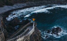From above back view of person with outstretched arms enjoying freedom standing on old stone bridge against troubled water with foam waves washing rocky shore in Spain — Stock Photo