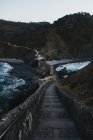 Leere Pflastersteine, die entlang einer Brücke zwischen Steinzäunen und an einer Seite eines felsigen Hügels entlang der Uferwäsche durch unruhiges Meerwasser mit weißen Schaumwellen in der Dämmerung in Spanien führen — Stockfoto