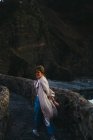 Женщина в повседневной одежде ходит по старому каменному мосту улыбаясь и глядя в камеру на проблемную заливную воду, моющую скалистое побережье Испании — стоковое фото