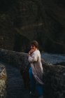 Angle élevé de la femelle en vêtements décontractés avec grand chien brun debout sur un vieux pont de pierre appuyé sur la clôture et regardant loin avec intérêt contre l'eau de baie troublée laver la côte rocheuse en Espagne — Photo de stock
