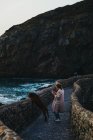 High angle of female in casual clothes with big brown dog standing on old stone bridge leaning on fence and looking away with interest against troubled bay water washing rocky coast in Spain — Stock Photo