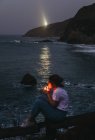 Da sopra vista laterale di casuale pensoso giovane sigaretta femminile fumare mentre seduto sulla recinzione sulla costa rocciosa del mare in estate sera nella città di Lekeitio in Spagna con luce faro sullo sfondo — Foto stock