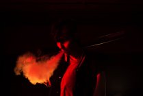Современная женщина в бронежилете с чёрной бейсбольной битой на плече во время курения с красным светом на заднем плане — стоковое фото