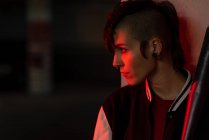 Side view of contemporary female in bomber jacket looking away while leaning on wall with black baseball bat on shoulder with red light on background — Stock Photo
