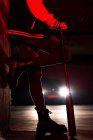 Боковой вид обрезанных ног безликой женщины в джинсах, опирающейся на стену и черные сапоги с массивной подошвой, держащей летучую мышь с автомобильными фарами на заднем плане — стоковое фото