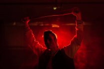 Femme rebelle en veste décontractée avec piercing et coiffure moderne tenant chauve-souris parmi la lumière rouge colorée et la vapeur — Photo de stock