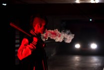Seitenansicht einer zeitgenössischen Frau in Bomberjacke mit einem schwarzen Baseballschläger auf der Schulter beim Rauchen mit rotem Licht und Autoscheinwerfern im Hintergrund — Stockfoto