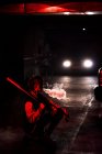 Вид сбоку на неузнаваемую современную женщину, присевшую в пиджаке бомбардировщика с чёрной бейсбольной битой на плече во время курения с красным светом и автомобильными фонарями на заднем плане — стоковое фото