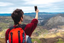 Back view of unrecognizable woman with backpack standing with arm raised and taking shot with smartphone of wonderful scenery with small villages and town in valley against foggy ridges at horizon under cloudy sky in Asturias — Stock Photo