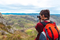 Side view of woman with backpack standing on glade and taking shot with camera of magnificent valley against foggy ridges at horizon under sky with fluffy clouds in Spain — Stock Photo