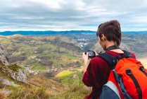 З боку жінка з рюкзаком стоїть на галявині і знімає з фотоапаратом величну долину проти туманних кряжів під небом з пухнастими хмарами в Іспанії. — стокове фото
