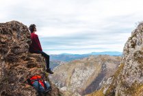 Vista lateral del turista sentado en el borde del acantilado disfrutando de la libertad y admirando increíbles paisajes de campo ubicados en el valle al pie de la montaña contra colinas boscosas brumosas y llanura bajo el cielo con nubes grises exuberantes en España - foto de stock