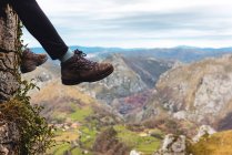 Vue latérale des jambes touriste assis sur le bord de la falaise jouissant de la liberté et admirant les paysages étonnants de la campagne située dans la vallée au pied de la montagne contre les collines boisées brumeuses et la plaine sous le ciel avec des nuages gris luxuriants en Espagne — Photo de stock