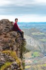 Vue latérale du touriste assis sur le bord de la falaise jouissant de la liberté et admirant les paysages étonnants de la campagne située dans la vallée au pied de la montagne contre les collines boisées brumeuses et la plaine sous le ciel avec des nuages gris luxuriants en Espagne — Photo de stock