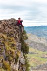 Вид сбоку на туриста, сидящего на краю скалы, наслаждающегося свободой и восхищающегося удивительной живописностью сельской местности, расположенной у подножия горы на фоне туманных холмов и равнины под небом с пышными облаками в Испании — стоковое фото