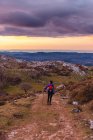Back view of tourist with backpack and stick looking away and admiring picturesque scenery while hiking on hill road under cloudy sky in Spain — Stock Photo