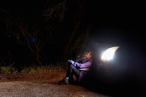 Provocativa mujer confiada en violeta y vibrante chaqueta naranja iluminación cigarrillo mientras se sienta solo apoyado en el automóvil con faros brillantes en la noche oscura en España - foto de stock