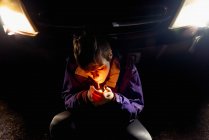 D'en haut provocateur confiant femelle en violet et veste orange vif allumant cigarette tout en étant assis seul appuyé sur l'automobile avec des phares lumineux dans la nuit noire en Espagne — Photo de stock