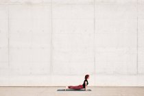 Vista lateral del atleta femenino de moda en ropa deportiva haciendo ejercicio de yoga cobra bebé en alfombra deportiva mientras entrena solo en la calle - foto de stock