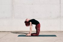 Trendy fit Frau in Sportbekleidung macht Yoga in Kamelpose auf Sportmatte allein auf der Straße gegen Betonwand bei sonnigem Tag — Stockfoto