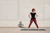 Ajustez femme réfléchie avec des cheveux teints en rouge bouclé dans les vêtements de sport contemplant tout en se tenant debout sur le tapis de yoga contre le mur de béton — Photo de stock