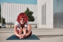 Flessibile atleta donna scalza con i capelli rossi ricci in activewear seduto in paschimottanasana e sorridente durante la pratica dello yoga su strada — Foto stock