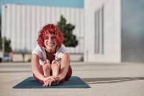 Deportista femenina descalza flexible con el pelo rizado rojo en ropa deportiva sentada en paschimottanasana y sonriendo mientras practica yoga en la calle - foto de stock