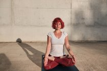 Donna con capelli rossi e tatuaggio in camicia bianca e pantaloni bordeaux seduta in padmasana con Gyan Mudra e meditando ad occhi chiusi — Foto stock