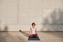Woman with red hair and tattoo in white shirt and burgundy pants sitting in padmasana with Gyan Mudra and meditating with closed eyes — Stock Photo