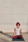 Женщина с рыжими волосами и татуировкой в белой рубашке и бордовыми брюками сидит в падмасане с Гьяном Мудрой и медитирует с закрытыми глазами — стоковое фото