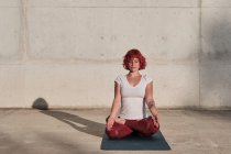 Woman with red hair and tattoo in white shirt and burgundy pants sitting in padmasana with Gyan Mudra and meditating with closed eyes — Stock Photo