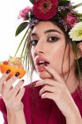Молода розкішна жінка з вінком з різнокольорових квітів на голові і пальці на губі дивиться на камеру, тримаючи тарілку в помаранчевій цеглинці ізольовано на білому тлі — стокове фото