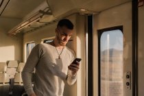 Joven enfocado en auriculares inalámbricos mensajería de texto en el teléfono móvil mientras está de pie y apoyado en la pared del tren a la luz del sol - foto de stock
