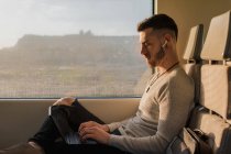Вид збоку молодого чоловіка в бездротових навушниках, що друкуються на ноутбуці під час подорожі в метро в сонячний день — стокове фото