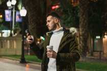 Konzentrierter junger Mann mit Imbissbecher-SMS auf dem Handy, während er abends auf der Straße steht — Stockfoto
