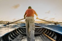 Back view of faceless man in casual wear standing in wooden boat holding big oars in hands with golden sunset on background — Stock Photo
