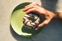 De cima colheita fêmea segurando donut gostoso com cobertura acima placa de cerâmica verde, tendo café da manhã no terraço do café em dia ensolarado — Fotografia de Stock