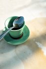 Сверху зеленая керамическая чашка теплого черного кофе проливается на стол, состоящий из металлической чайной ложки на блюдце на сером столе — стоковое фото