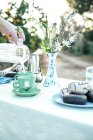 Senhora da colheita derramando leite de jarro transparente para copo verde ao fazer café à mesa com flores silvestres em vaso e rosquinhas frescas no prato descansando na natureza — Fotografia de Stock