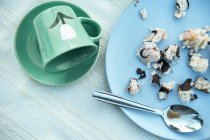 Green ceramic mug dropped on saucer and blue round plate with small crumbs of tasty pastry in composition with dirty metal teaspoon after breakfast — Stock Photo