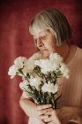 De cima velha fêmea pensativa com cabelo grisalho com olhos fechados segurando buquê de cravo branco em casa — Fotografia de Stock