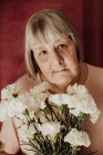 Зверху вдумлива стара жінка з сірим волоссям дивиться на камеру і тримає букет білої гвоздики вдома — стокове фото