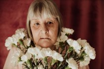 From above thoughtful old female with gray hair looking at camera holding bouquet of white carnation at home — Stock Photo