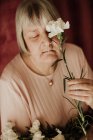 Von oben eine nachdenkliche alte Frau mit grauen Haaren und geschlossenen Augen, die zu Hause einen Strauß weißer Nelken hält — Stockfoto