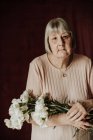 From above thoughtful old female with gray hair looking at camera and holding bouquet of white carnation at home — Stock Photo