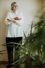 Сосредоточенная пенсионерка в белой блузке и черных брюках, стоящих со скрещенными руками на стене и смотрящих в сторону — стоковое фото