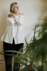 Зосереджена жінка-пенсіонерка у білій блузці та чорних штанах, що стоять з схрещеними руками, дивлячись на стіну — стокове фото