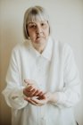 Triste donna anziana in camicetta bianca gesticolare con le mani su sfondo chiaro guardando la fotocamera — Foto stock