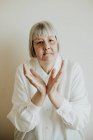 Traurige ältere Frau in weißer Bluse, die Uneinigkeit zeigt, während sie vor hellem Hintergrund die Hände hebt und in die Kamera blickt — Stockfoto